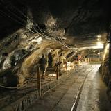 Korytarz z solnymi postaciami stojącymi wzdłuż szyn. W głębi korytarza więcej światła, a na pierwszym planie mrok w kopalni soli w Wieliczce.